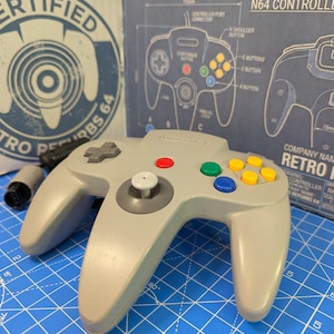 May include: A gray Nintendo 64 controller with colored buttons and a directional pad. The controller is on a blue grid surface, with a blueprint and a "CERTIFIED RETRO REFURBS 64" logo in the background. The controller has the Nintendo logo.