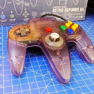 May include: A translucent purple video game controller featuring a gray directional pad, a white central analog stick, one red button, and four colored C-buttons in yellow, green, and blue. The controller is positioned in front of a box labeled Retro Refurbs 64.
