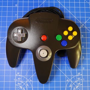 May include: A black Nintendo 64 video game controller featuring a grey directional pad, a central grey analogue stick, a red start button, a green B button, a blue A button, and four yellow C-buttons on the right side with a black cord attached.