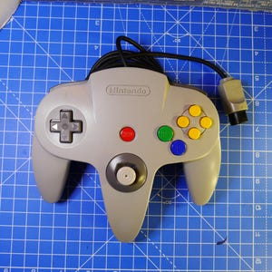 May include: A gray Nintendo 64 video game controller featuring a central analog stick, a directional pad on the left, and four colored action buttons on the right, including yellow, green, and blue, with a red start button and a black cord.