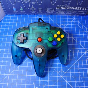 Puede incluir: Un mando de videojuegos de color azul verdoso translúcido con un joystick analógico gris central, una cruceta gris, un botón de inicio rojo y cuatro botones de acción de colores amarillo, verde y azul. El logotipo de Nintendo está grabado.