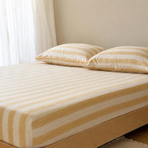 Könnte beinhalten: Ein Bett mit einem Spannbettlaken und zwei passenden Kopfkissen, die alle mit einem Muster aus breiten, horizontalen Streifen in abwechselnden Tönen von sanftem Goldgelb und Off-White bezogen sind. Die Bettwäsche hat eine Textur.