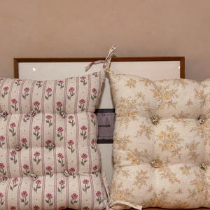 May include: Two square tufted seat cushions with fabric ties at the corners. The left cushion features a cream background with vertical pink stripes and small pink floral motifs. The right cushion has a cream background with a dense yellow floral pattern.