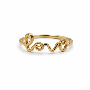 Può includere: Un anello color oro con la parola "love" scritta in corsivo. L'anello è realizzato in metallo lucido e liscio, su sfondo bianco. Un gioiello semplice ed elegante.