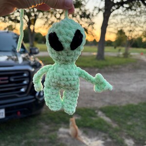 May include: A crocheted alien figure in light green with black eyes. The handmade plush toy has outstretched arms and is suspended by a matching green string. The alien is held up against a blurred outdoor background.