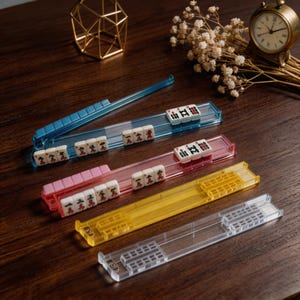 Könnte beinhalten: Vier farbige Mahjong-Kartenhalter in Blau, Rosa, Gelb und Transparent. Jeder Halter enthält eine Reihe weißer Spielsteine mit roten und schwarzen Markierungen. Die Halter sind aus transparentem Kunststoff gefertigt und dienen dazu, die Spielsteine während des Spiels zu halten.