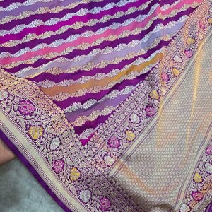 May include: A colourful textile with horizontal stripes in shades of purple, pink, yellow, and white. The border is decorated with floral and leaf patterns in gold, white, and pink. The fabric has a shimmering, luxurious appearance.