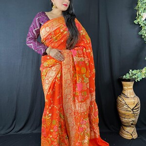 Puede incluir: Un sari naranja con estampados florales y un borde dorado. El sari está drapeado sobre una persona que lleva una blusa morada de manga larga. El fondo es una cortina oscura.