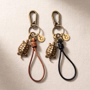 May include: Two keychains featuring antique brass-toned turtle charms, a circular tag engraved with the letter C, and a metal lobster clasp. One keychain has a tan leather loop, and the other has a black leather loop, both attached to a metal ring.