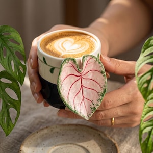 May include: A ceramic mug featuring a two-tone design with a cream-colored upper section and a dark blue lower section. The mug is adorned with a three-dimensional, sculpted pink leaf handle with prominent red veins and green speckled edges.