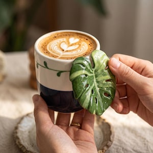 May include: A ceramic mug featuring a two-tone design with a white upper section and a dark blue lower section. The mug is adorned with a green, glossy, three-dimensional monstera leaf handle and a delicate hand-painted green vine pattern.