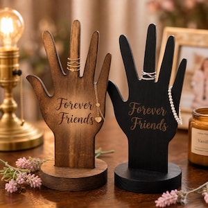 May include: Two wooden hand-shaped jewelry holders, one stained brown and the other black. The brown holder has a gold necklace and rings, while the black holder displays a pearl necklace and rings. Both have the text "Forever Friends".