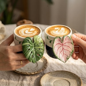 Puede incluir: Dos tazas de cerámica llenas de café, cada una con un asa tridimensional en forma de hoja. Una taza tiene un asa de hoja de monstera verde, mientras que la otra presenta un asa de hoja rosa y verde en forma de corazón. Ambas tazas sobre platillos.