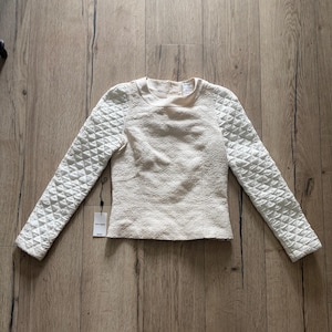 Könnte beinhalten: Cremefarbenes Langarm-Oberteil mit gesteppten Ärmeln und strukturiertem Körper. Das Oberteil hat einen Rundhalsausschnitt und ein angebrachtes Etikett. Die Ärmel weisen ein Rautenmuster auf, während der Körper ein subtiles, wirbelndes Design hat. Das Kleidungsstück wird auf einer Holzoberfläche präsentiert.