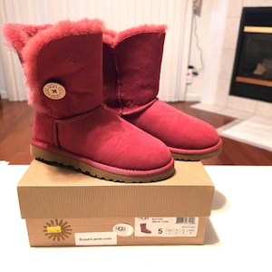 May include: A pair of mid-calf boots in a vibrant raspberry pink color, featuring a soft, plush interior lining and a single large wooden button on the side with the UGG logo. The boots are resting on a cardboard box with product labels.