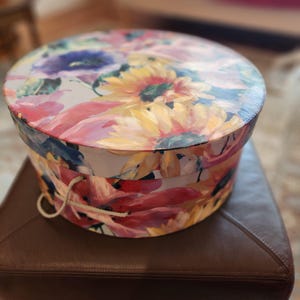 May include: A round hat box with a floral design featuring yellow sunflowers, pink and red petals, and blue accents. The box has a white background and a rope handle. The box is sitting on a brown leather ottoman.