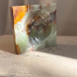 Può includere: Un oggetto decorativo in pietra di forma quadrata con una superficie lucida caratterizzata da bande traslucide color verde pallido, bianco e ambra scuro. La pietra presenta venature naturali e texture cristalline su tutta la forma piatta.