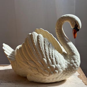 May include: A cream-coloured ceramic swan figurine with a glossy finish. The swan features detailed moulded feather textures on its wings and body, a curved neck, and a beak painted in shades of orange and black with a distinct black eye marking.