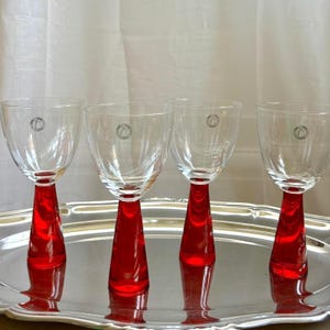 May include: Four clear wine glasses with vibrant red stems, resting on a silver tray. Each glass has a circular emblem near the top. The glasses are designed with a classic shape, suitable for serving wine.