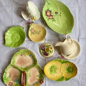 Puede incluir: Una colección de vajilla de cerámica que incluye un cuenco grande con forma de hoja verde, un plato pequeño de hoja verde, un plato de hoja amarilla, una bandeja de tres secciones de hoja rosa y verde, un plato floral amarillo, una jarra de crema con platillo y un portavelas floral.