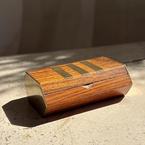 May include: A rectangular wooden box with a hinged lid featuring three parallel brass inlays across the top. The box has a polished wood grain finish and brass metal edges on the sides, with a small visible chip along the front lid seam.