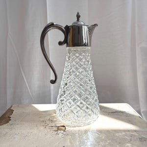 Könnte beinhalten: Ein klarer Glaskrug mit Rautenmuster, ausgestattet mit einem silberfarbenen Metalldeckel und einem geschwungenen Griff. Das Gefäß hat eine konische Form, einen spitzen Ausguss und einen kleinen Knauf auf dem Deckel mit metallischem Glanz.