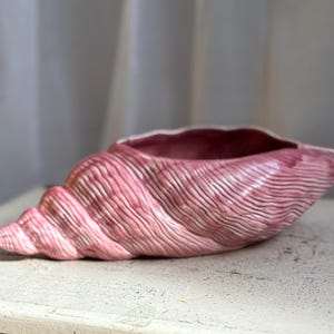 Op de afbeelding: Een keramische schelpvormige plantenbak met een glanzende roze afwerking en diepe, getextureerde ribbels aan de buitenkant. De binnenkant van de schelp is hol en glad, met een bijpassend roze glazuur over het gehele decoratieve object.