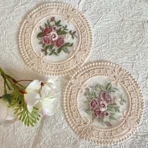 May include: Two round lace coasters featuring a cream-coloured crocheted border and a central sheer panel embroidered with a floral motif in shades of dusty rose, dark pink, and sage green, accompanied by a cluster of white artificial flowers.