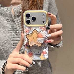 May include: A clear phone case with a yellow border and a cartoon design. The case features a star-shaped pop socket with a cartoon character and a blue background with stars and other characters. The phone case has the word "NOVA" printed on it.