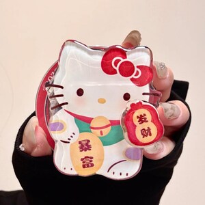 May include: Clear acrylic charm featuring a cartoon cat with a red bow, holding a red bag with Chinese characters. The charm has a red border and a gold bell. The Chinese characters on the charm read "Wealth".