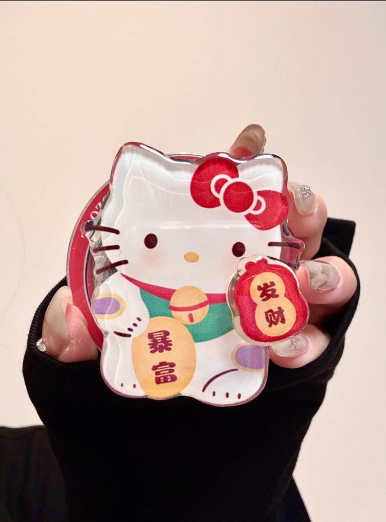 May include: Clear acrylic charm featuring a cartoon cat with a red bow, holding a red bag with Chinese characters. The charm has a red border and a gold bell. The Chinese characters on the charm read "Wealth".