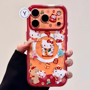 May include: A clear phone case with a red border featuring a design with Hello Kitty characters. The case has an orange background with various Hello Kitty figures, flowers, and other decorative elements. The case has cutouts for the phone's camera.