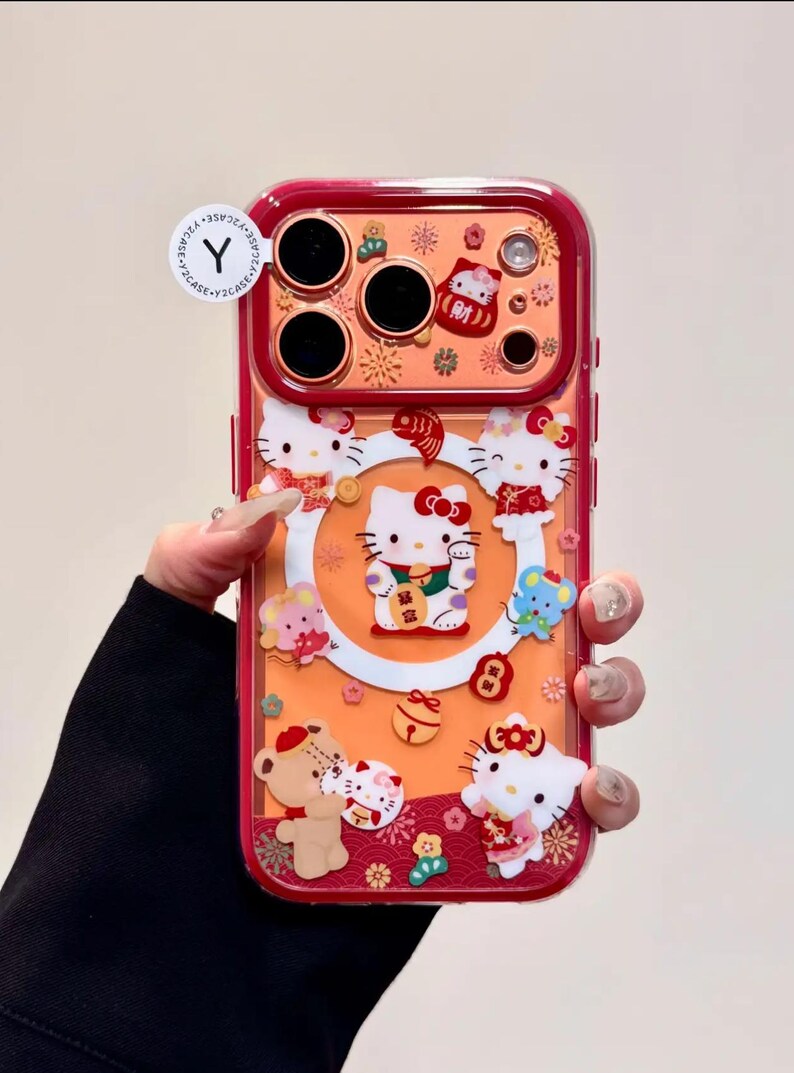 May include: A clear phone case with a red border featuring a design with Hello Kitty characters. The case has an orange background with various Hello Kitty figures, flowers, and other decorative elements. The case has cutouts for the phone's camera.