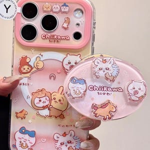 May include: A pink and clear phone case with a matching pop socket. The case and pop socket feature cartoon characters and the Japanese text "Chiikawa". The case has a pink border and cute illustrations.