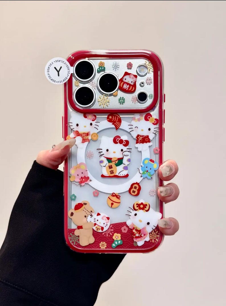 May include: A clear phone case with a red border, featuring multiple Hello Kitty figures and other decorative elements. The case has a festive design with a central Hello Kitty figure holding a gold ingot. The case has a Y2CASE sticker.