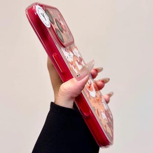 May include: A red phone case with a clear back featuring a cartoon design. The case has a glossy finish and a raised edge for screen protection. The design includes various cartoon characters in shades of brown, white, and red.