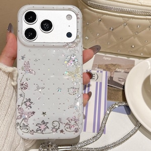 May include: A white smartphone case featuring a subtle polka dot pattern, iridescent butterfly and floral motifs, and small star accents. The case includes a silver-toned metal camera frame and a detachable, shimmering rhinestone-encrusted wrist strap.