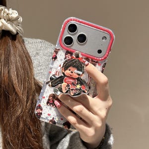 May include: Clear phone case with a red border and a cartoon character design. The case features a pop-up grip with a matching character. The case has a pattern of small cartoon characters and hearts.