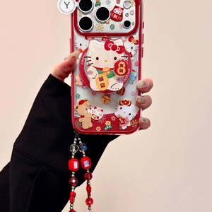 May include: Red and clear phone case featuring Hello Kitty and other cartoon characters. The case has a red border and a ring holder. A beaded lanyard with red and clear beads is attached to the case.
