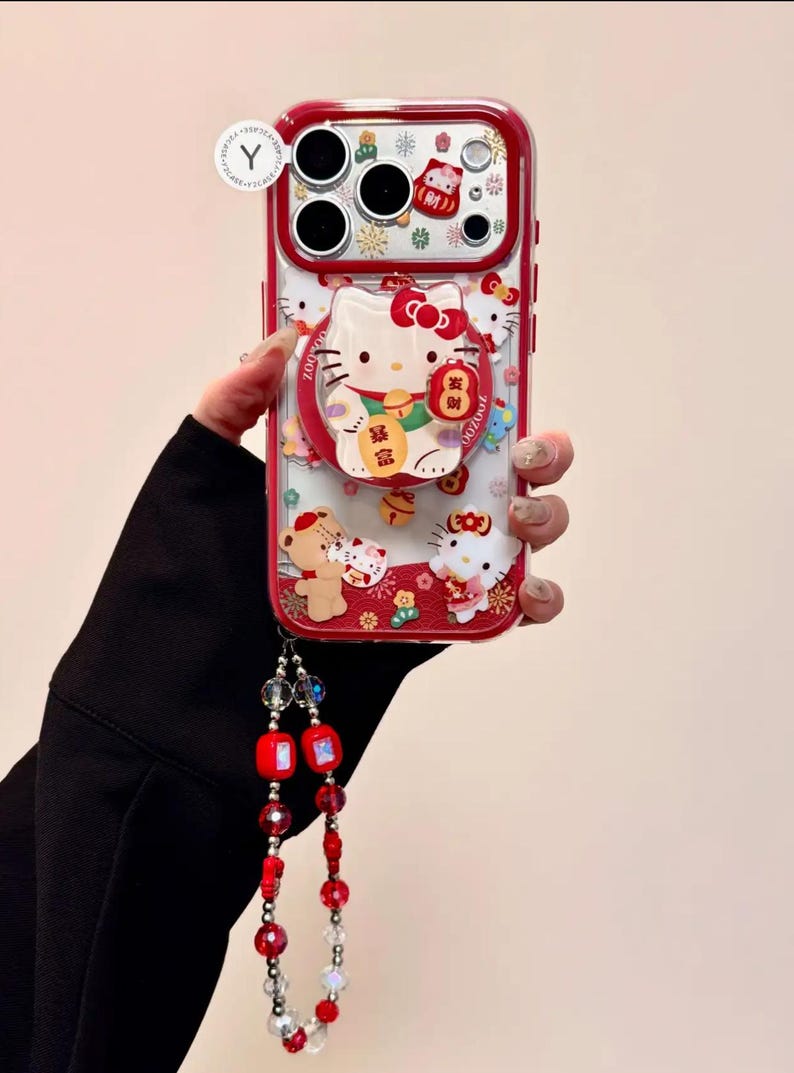 May include: Red and clear phone case featuring Hello Kitty and other cartoon characters. The case has a red border and a ring holder. A beaded lanyard with red and clear beads is attached to the case.