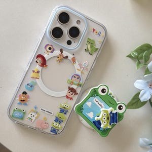 May include: A clear phone case featuring various colorful cartoon character stickers, including a green dinosaur, a space ranger, a cowboy, and aliens. The case has a white circular magnetic ring and the text Keep childish. A green frog phone grip is nearby.