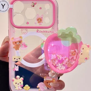 May include: A pink and clear phone case featuring Rilakkuma bear characters. The case has a strawberry-shaped charm filled with small bear figures. The design includes the text "Rilakkuma" and Japanese text. The case is designed for a phone.
