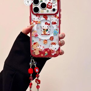 May include: A red and clear phone case featuring Hello Kitty and other cartoon characters. The case has a red border and a decorative wrist strap with red beads. The design includes festive elements.