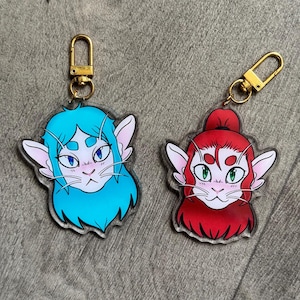 Pode incluir: Dois chaveiros de acrílico com personagens de desenho animado semelhantes a gatos. Um tem cabelo azul e o outro vermelho. Ambos têm fechos dourados. Os chaveiros estão em uma superfície de madeira.