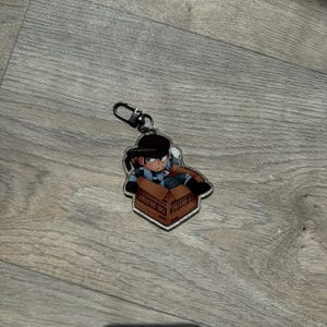 May include: An acrylic keyring featuring a cartoon character with dark hair and a headband, wearing a blue and black tactical outfit, sitting inside a brown cardboard box with the text THE ORANGE printed on the side, attached to a black metal clasp.