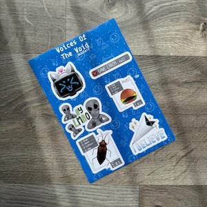 May include: A blue sticker sheet with various designs, including a robot cat, aliens, a burger, and the text "I BELIEVE." The sheet also features the text "Voices Of The Void Stickers."