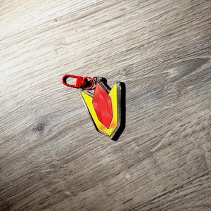 May include: A keychain featuring a shield-shaped design with a red gem at the center. The shield has yellow and orange gradients, and a red clasp. The keychain is made of clear acrylic.