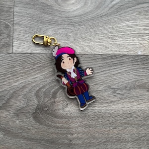 May include: A colorful acrylic keychain featuring a cartoon character in a Renaissance-style outfit. The character has brown hair, a pink and blue hat, and is holding a lute. The keychain has a gold-colored clasp.
