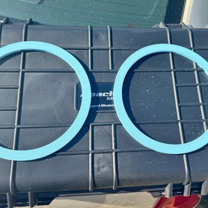Puede incluir: Dos juntas circulares de color azul claro se muestran sobre una maleta negra texturizada. La maleta tiene un patrón de rejilla y el texto "pact" es visible. Las juntas están espaciadas uniformemente y parecen estar hechas de un material flexible.