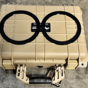 Puede incluir: Una maleta rectangular de color beige con un asa y dos inserciones circulares negras. La maleta tiene una superficie texturizada y cierres metálicos. La marca "Alpine" es visible en la maleta.